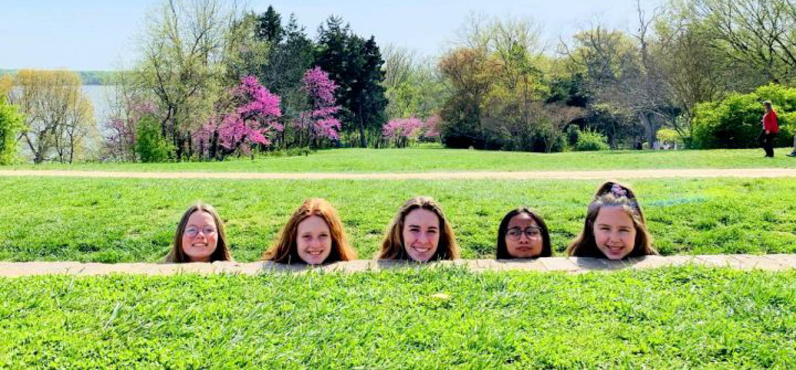 Five female students with only their heads showing above a grassy ledge